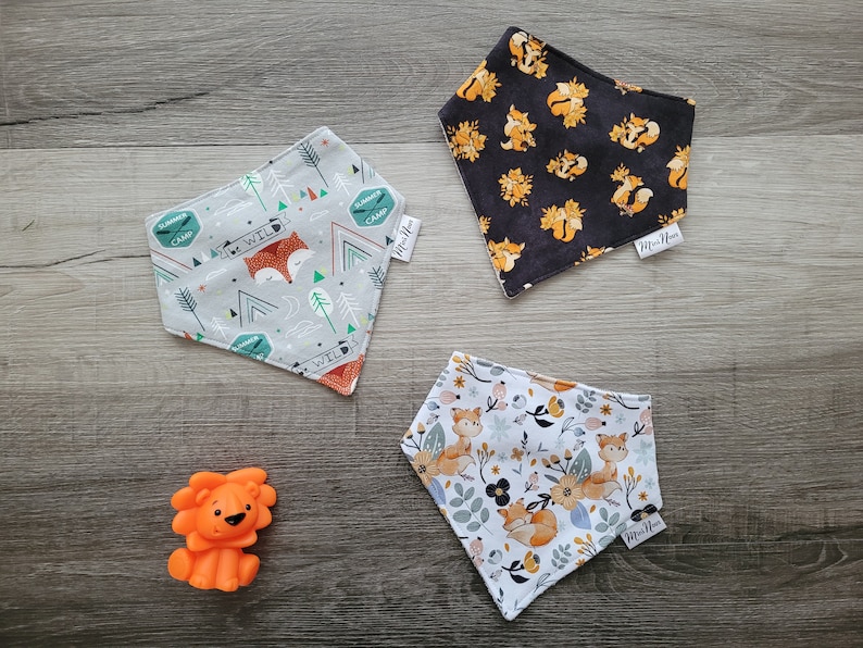 Puede incluir: Tres baberos bandana con tem&aacute;tica de zorros con diferentes patrones. Un babero es gris con acentos verdes y naranjas y el texto "Be Wild". Otro babero es negro con zorros naranjas. El tercer babero es blanco con zorros naranjas y acentos florales. Todos los baberos tienen el texto "Mini-Me" en la esquina inferior derecha.