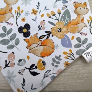 Puede incluir: Tela blanca con un patr&oacute;n repetido de zorros naranjas, flores amarillas y hojas grises. La tela tiene una etiqueta blanca con el texto "Mini Nous" en negro.