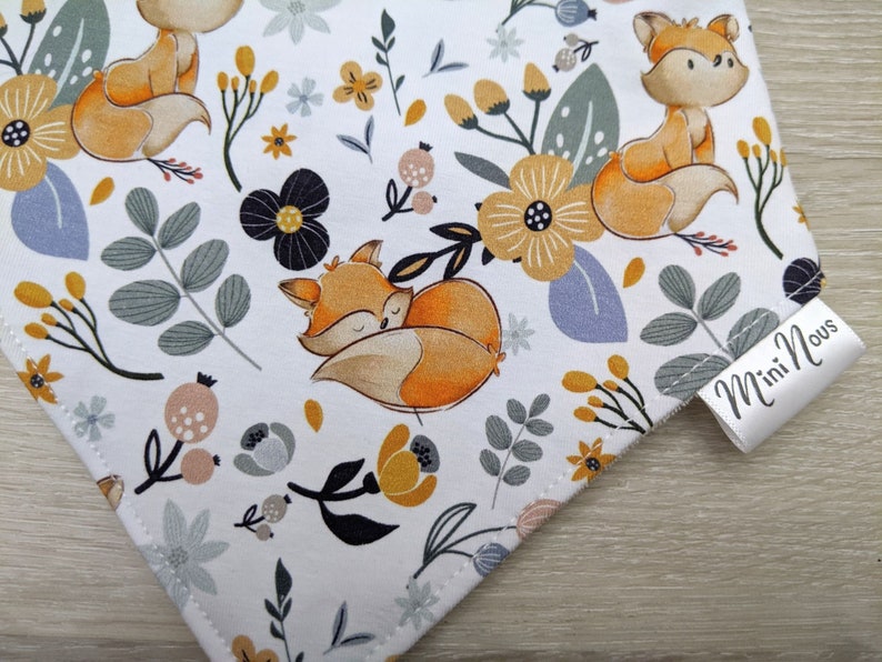 Puede incluir: Tela blanca con un patr&oacute;n repetido de zorros naranjas, flores amarillas y hojas grises. La tela tiene una etiqueta blanca con el texto "Mini Nous" en negro.