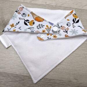 Puede incluir: Babero blanco para beb&eacute; con un patr&oacute;n floral en tonos amarillos, grises y naranjas. El babero tiene dos broches blancos.