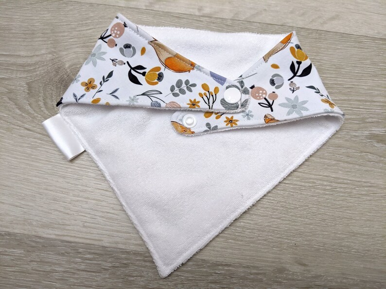 Puede incluir: Babero blanco para beb&eacute; con un patr&oacute;n floral en tonos amarillos, grises y naranjas. El babero tiene dos broches blancos.