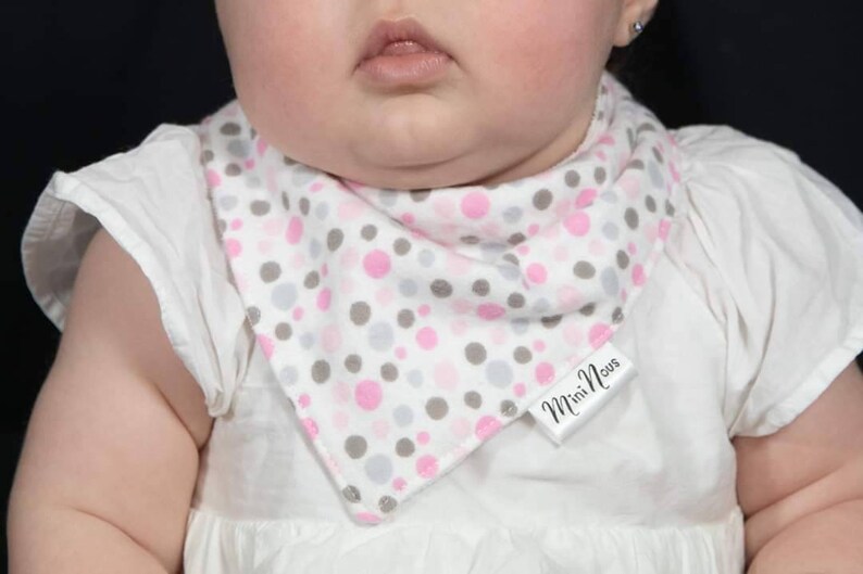 Puede incluir: Un babero blanco para beb&eacute; con un patr&oacute;n de lunares rosa y gris. El babero tiene una etiqueta blanca con el texto "Mini Nous" en &eacute;l.