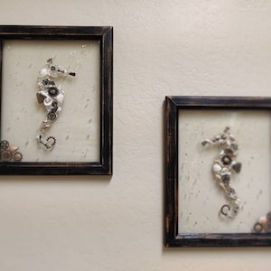 May include: Two framed pieces of art featuring seahorses made from shells and other found objects. The seahorses are white and the frames are black.