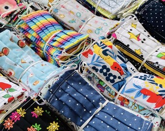 May include: A collection of assorted fabric face masks, each folded and secured with white elastic. The masks display a variety of patterns, including polka dots, floral designs, and rainbow stripes. The masks are in various colors, including blue, black, and yellow.