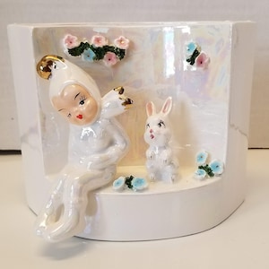 Könnte beinhalten: Weißer Keramik-Blumentopf mit einem fantasievollen Design, das eine feenhafte Figur mit einer goldenen Krone und ein weißes Kaninchen zeigt. Der Topf hat einen gewellten Rand und ist mit blauen und rosa Blumen verziert.