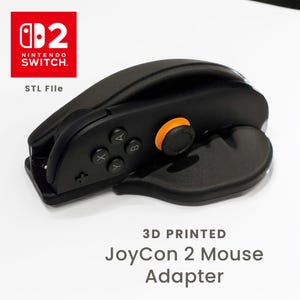 以下が含まれることがあります： Nintendo Switch用の黒い3DプリントJoyCon 2マウスアダプター。アダプターにはオレンジ色のボタンと、A、B、X、Yとラベル付けされた黒いボタンがあります。Nintendo Switchのロゴは左上にあり、その下に「STL File」というテキストがあります。