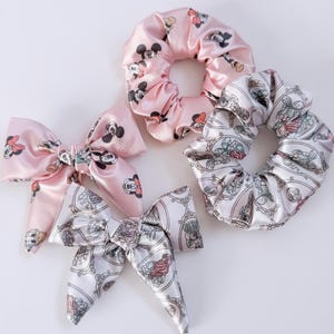 May include: A collection of satin hair accessories, including two scrunchies and two bow-tie hair clips. One scrunchie and one bow are light pink with a Mickey Mouse pattern. The other scrunchie and bow are white with a princess design.