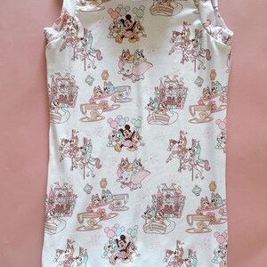 May include: A sleeveless white romper featuring a repeating pattern of cartoon characters including Bluey, Bingo, Mickey Mouse, and Dumbo. The print includes illustrations of amusement park rides like teacups, carousels, and a castle in soft pastel colors.