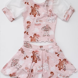 以下が含まれることがあります： 女の子用のピンクのツーピースの服。半袖のトップスとスカート。どちらもトイストーリーのキャラクター、ウッディ、バズ・ライトイヤー、ジェシー、ブルズアイがプリントされています。トップスは袖に白いシアーオーバーレイが付いています。