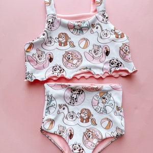 May include: A two-piece swimsuit with a repeating pattern of cartoon dogs, beach balls, and inflatable floats in light blue and pink. The top has a ruffled edge and straps, and the high-waisted bottoms complete the set. A fun and colourful design.