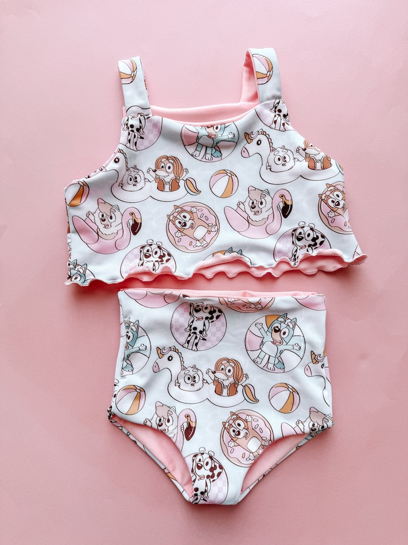 May include: A two-piece swimsuit with a repeating pattern of cartoon dogs, beach balls, and inflatable floats in light blue and pink. The top has a ruffled edge and straps, and the high-waisted bottoms complete the set. A fun and colourful design.