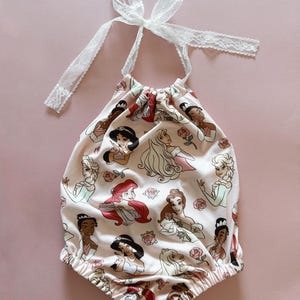 May include: A light pink baby romper featuring a pattern of cartoon princesses and roses. The romper has a halter-style neckline with a white lace tie. The bottom of the romper has elastic around the legs.