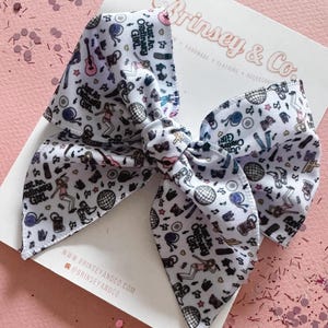 May include: A large white hair bow with a repeating pattern of musical instruments, disco balls, and dancing figures. The bow has the text "Just An Ordinary Girl" and the brand name "Brinsey & Co" printed on it.