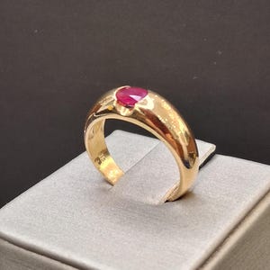 May include: A gold ring featuring a smooth, polished band and a single, oval-cut, deep red gemstone set securely in the center. The metal has a warm, reflective finish, and the interior of the band shows a small, stamped hallmark near the base.