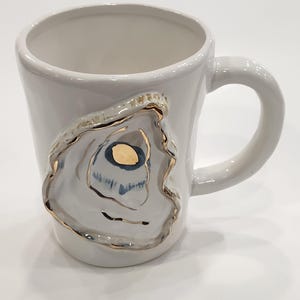 May include: White ceramic mug with a sculpted oyster shell design. The oyster shell has gold and blue accents, adding a touch of elegance. The mug has a curved handle and a smooth, glossy finish.