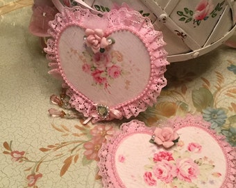 Puede incluir: Dos adornos en forma de corazón con ribete de encaje rosa y diseños florales. Cada corazón presenta una imagen central de rosas rosas, adornada con perlas, una pequeña rosa y una joya decorativa. Se colocan sobre una superficie con estampado floral.