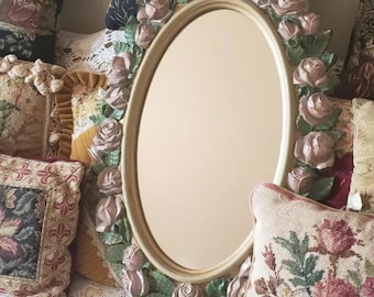 May include: An oval-shaped wall mirror featuring a cream-colored frame adorned with sculpted, light pink roses and green leaves encircling the entire perimeter of the glass surface. The mirror has a smooth, reflective center and a raised floral border.