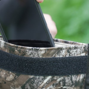 May include: A camouflage-patterned phone case with a black velcro strap. A black smartphone is being inserted into the case.