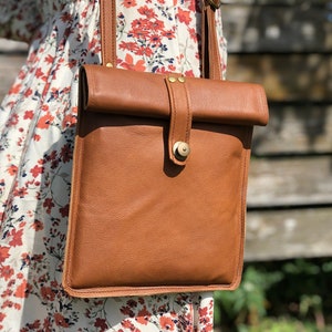 May include: A brown leather crossbody bag with a flap closure and a brass button. The bag is worn over the shoulder of a person wearing a floral dress.