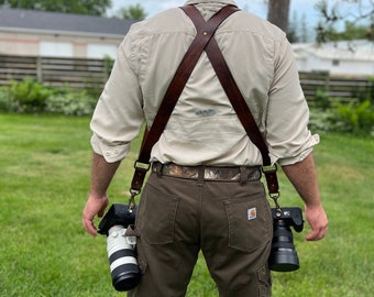 May include: A person wearing a brown leather camera strap with two cameras attached. The strap is worn over the shoulders and crosses at the back. The person is standing in a grassy area with a building in the background.