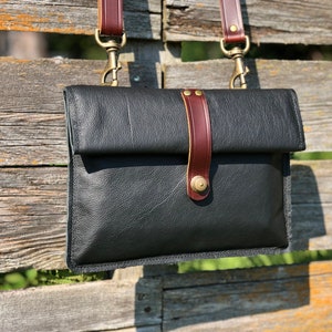 Könnte beinhalten: Schwarze Leder-Umhängetasche mit Klappenverschluss und braunem Lederriemen. Die Tasche hat messingfarbene Hardware und einen vertikalen braunen Lederakzentriemen mit Messingknopfverschluss. Die Tasche hängt an einem Holzzaun.