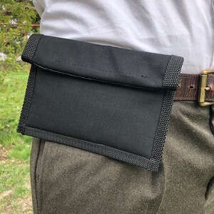 May include: A black fabric pouch with a zipper closure attached to a brown leather belt. The pouch is worn on the side of the waist.