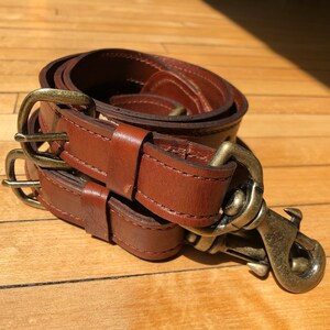 May include: A brown leather strap with two brass buckles and a brass hook. The strap is rolled up and the hook is visible.