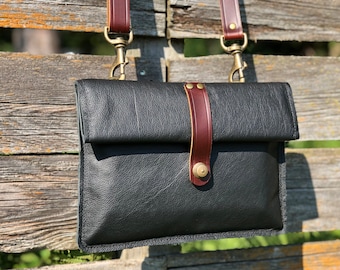 May include: Black leather crossbody bag with a flap closure and a brown leather strap. The bag features brass-colored hardware and a vertical brown leather accent strap with a brass button closure. The bag is hanging on a wooden fence.