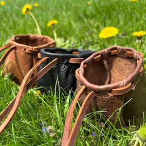 May include: Three small leather pouches with drawstrings, two brown and one black, are displayed on a bed of green grass. The pouches are handmade and have a rustic, vintage look.