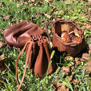 May include: Three brown leather pouches with drawstrings. Two of the pouches are open and one is closed. The open pouches contain small brown objects.