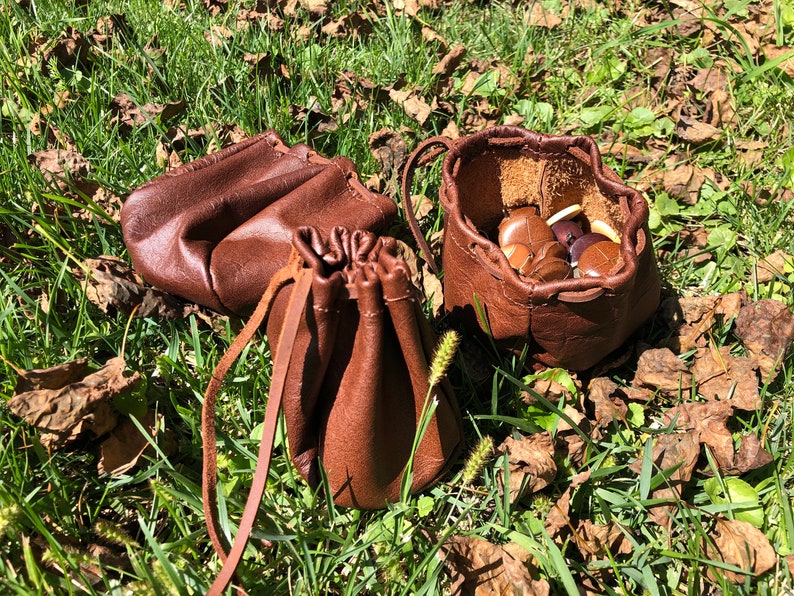 May include: Three brown leather pouches with drawstrings. Two of the pouches are open and one is closed. The open pouches contain small brown objects.