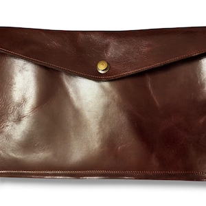 May include: A dark brown leather envelope-style clutch with a flap closure. The clutch has a shiny, polished finish and a gold-colored button clasp. The leather appears to be smooth and has a slight sheen, suggesting a high-quality material.
