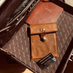 May include: A brown leather briefcase with a brown leather journal and a brown leather phone case inside. The phone case has a brown leather strap with a metal buckle. The briefcase has a brown fabric lining with a repeating pattern of the word "Wilson". The phone case has a business card inside that reads "Operation Genesis Gem Pouch - Genesis Passcard Pouch - William Passcard"