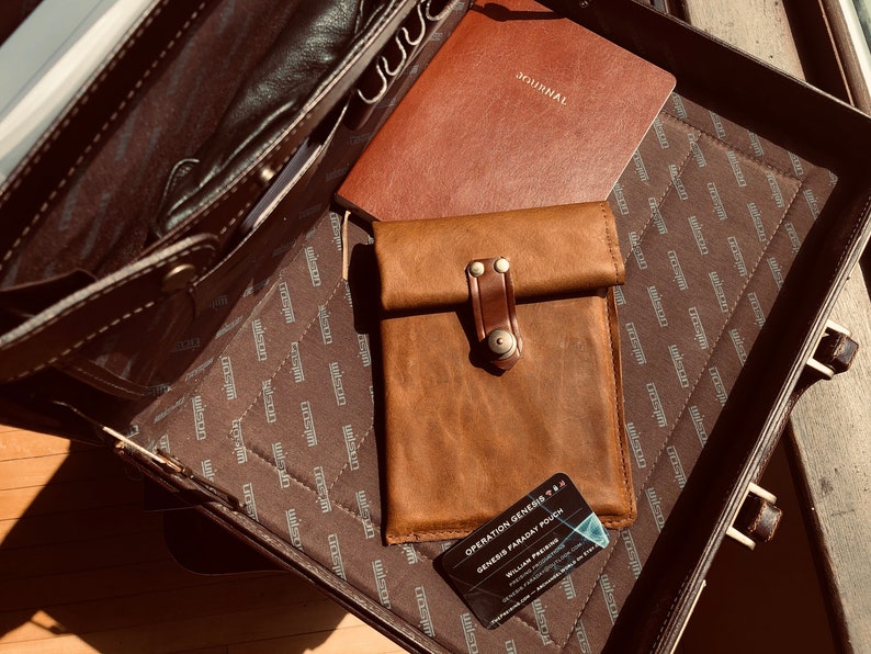 May include: A brown leather briefcase with a brown leather journal and a brown leather phone case inside. The phone case has a brown leather strap with a metal buckle. The briefcase has a brown fabric lining with a repeating pattern of the word "Wilson". The phone case has a business card inside that reads "Operation Genesis Gem Pouch - Genesis Passcard Pouch - William Passcard"