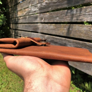 May include: A brown leather wallet with a brass snap closure. The wallet is folded and held in a hand.