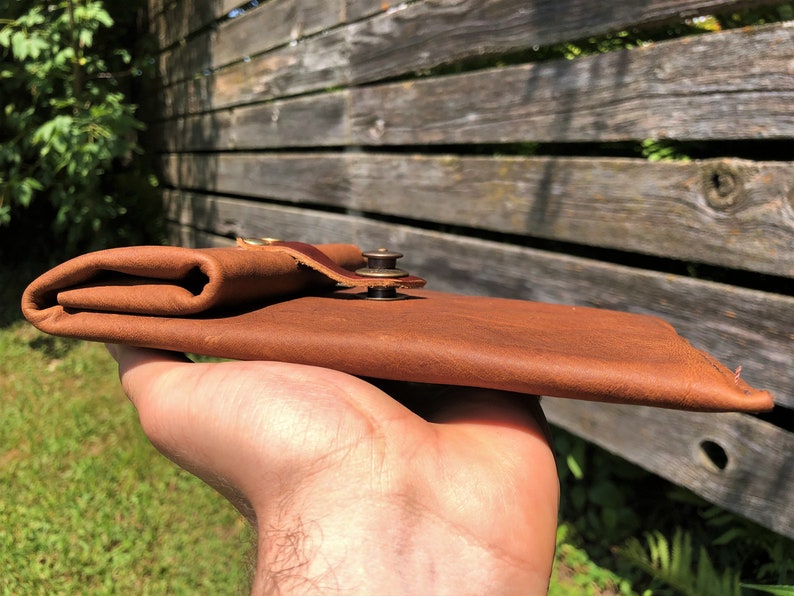 May include: A brown leather wallet with a brass snap closure. The wallet is folded and held in a hand.