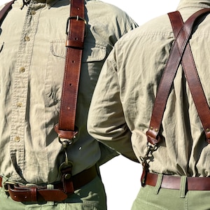 以下が含まれることがあります： 真鍮製の金具と調節可能なストラップが付いた茶色の革製のサスペンダー。サスペンダーは、薄い茶色のボタンダウンシャツと緑色のパンツの上に着用されています。