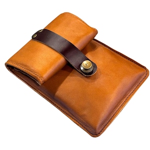 May include: A handcrafted, tan leather wallet with a dark brown strap and brass button closure. The wallet is rectangular with rounded corners and a stitched border. The design suggests a classic, durable accessory.