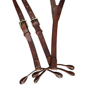 May include: Dark brown leather suspenders featuring adjustable straps with brass-toned metal buckles and reinforced stitching. The suspenders include four leather button-tab ends for attachment, showcasing a smooth finish and durable construction.