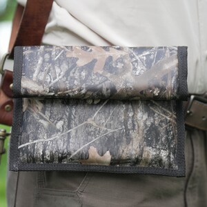 May include: A camouflage-patterned pouch with a black strap is worn on a person's belt. The pouch has a flap that folds over and is secured with a snap.