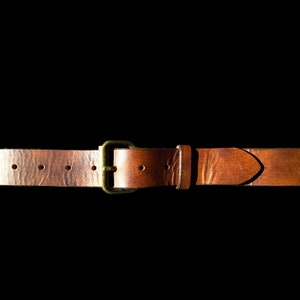 May include: A brown leather belt with a brass buckle. The belt has several holes for adjustment and a pointed end. The leather has a textured appearance and is set against a black background.