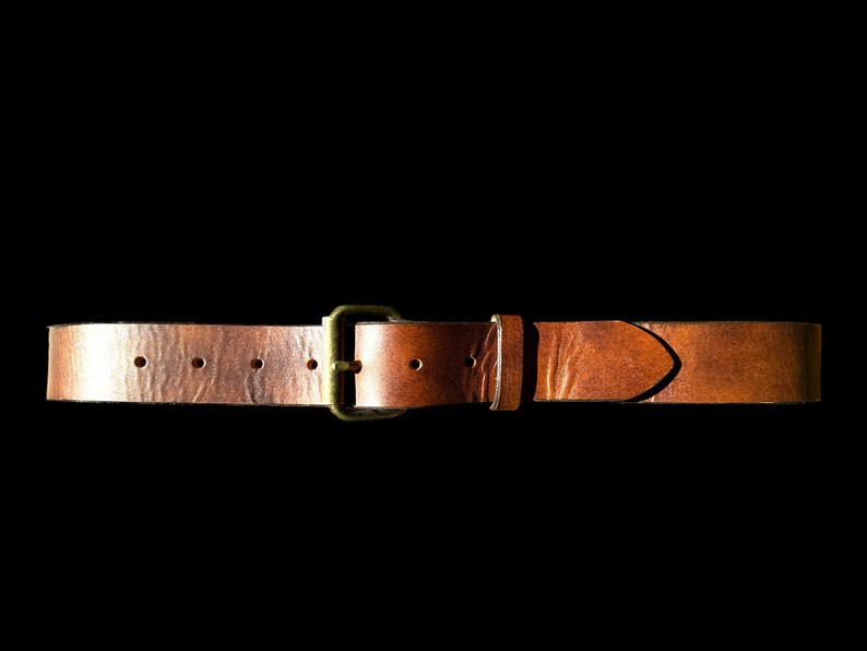 May include: A brown leather belt with a brass buckle. The belt has several holes for adjustment and a pointed end. The leather has a textured appearance and is set against a black background.