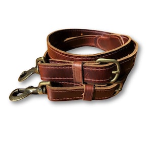 May include: Two brown leather straps with brass buckles and metal clasps. The straps are folded and stacked on top of each other.