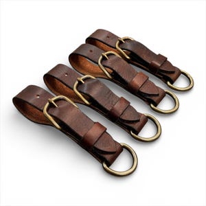 May include: Four brown leather keychains with brass buckles and rings. The keychains are arranged diagonally, showcasing the rich color and texture of the leather. Each keychain has a loop for keys.