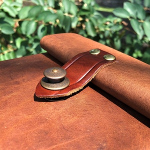 May include: Close-up of a brown leather journal with a brown leather strap and brass buckle. The strap is secured with two brass rivets.