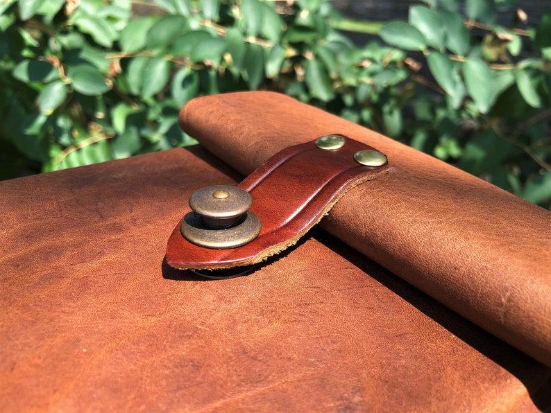 May include: Close-up of a brown leather journal with a brown leather strap and brass buckle. The strap is secured with two brass rivets.