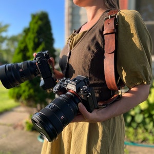 May include: A person wearing a brown leather camera strap with two black cameras attached. The cameras have large lenses and are positioned on the front of the strap.