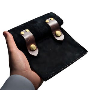 Puede incluir: Una cartera de cuero negro con correas de cuero marrón y remaches dorados. La cartera se sostiene en una mano, mostrando su diseño y artesanía. La cartera parece estar hecha de materiales de alta calidad.