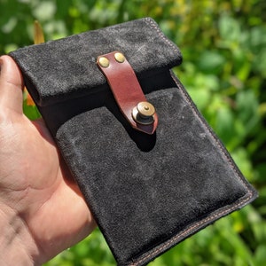 May include: A black suede phone case with a brown leather flap and a brass button closure. The case has a red stitch detail around the edges.