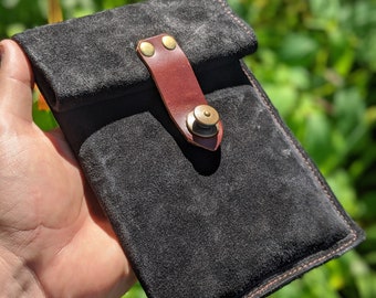 May include: A black suede phone case with a brown leather flap and a brass button closure. The case has a red stitch detail around the edges.